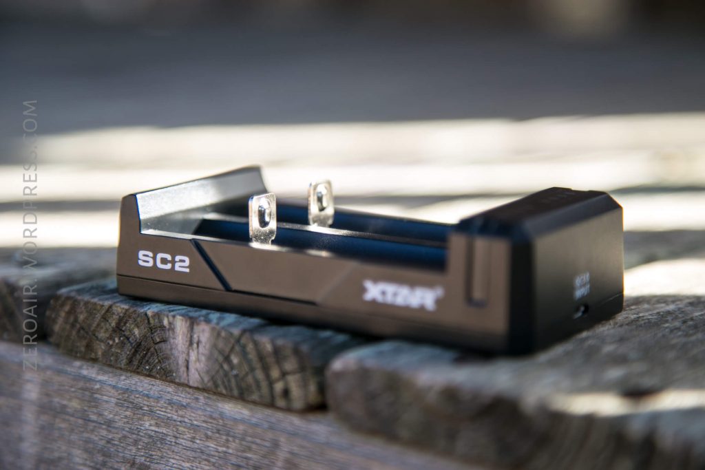 A black XTAR SC2 battery charger with two charging slots is placed on a weathered wooden surface outdoors, with sunlight casting soft shadows.