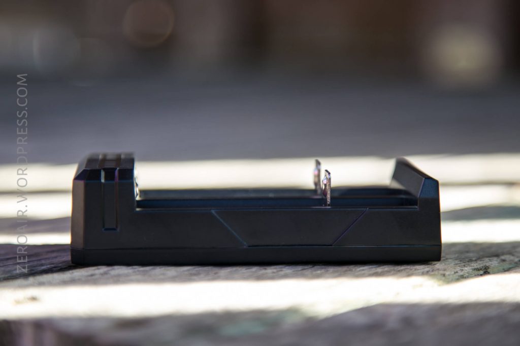 A black rectangular battery charger sits on a wooden surface with two metal contacts visible. Soft sunlight creates a blurred background. The words “ZER0NR.WORDPRESS.COM” are faintly visible along the left side.