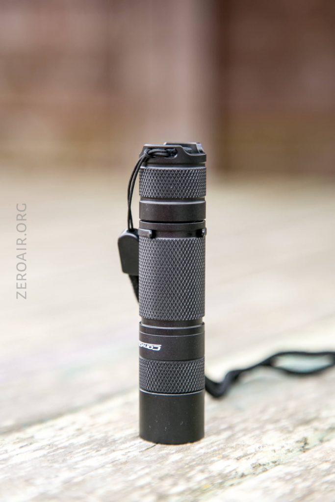 A small, black, textured flashlight stands upright on a wooden surface with a wrist strap attached. The background is blurred.