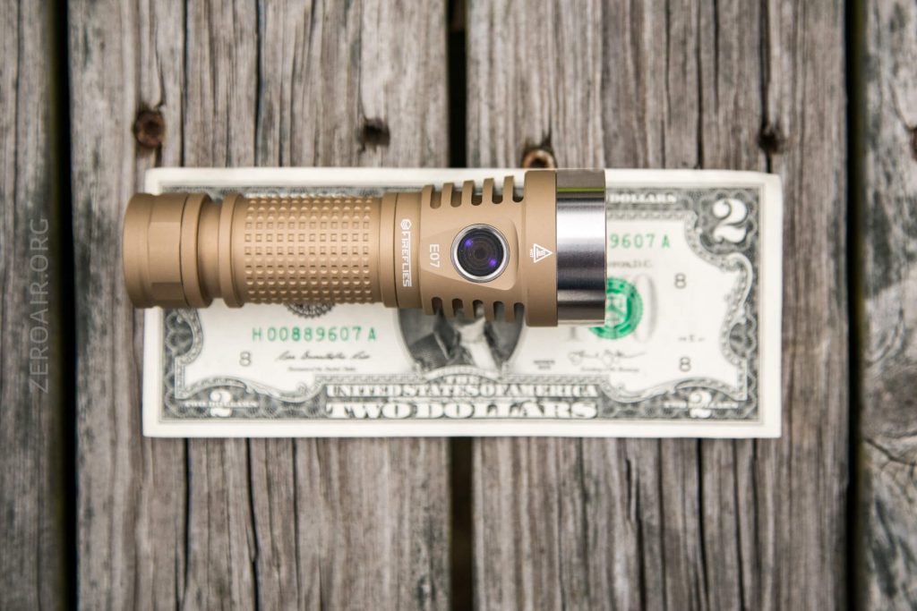 A bronze-colored flashlight is placed horizontally on top of a U.S. two-dollar bill, both resting on weathered wooden planks. The flashlight is slightly shorter than the length of the bill.