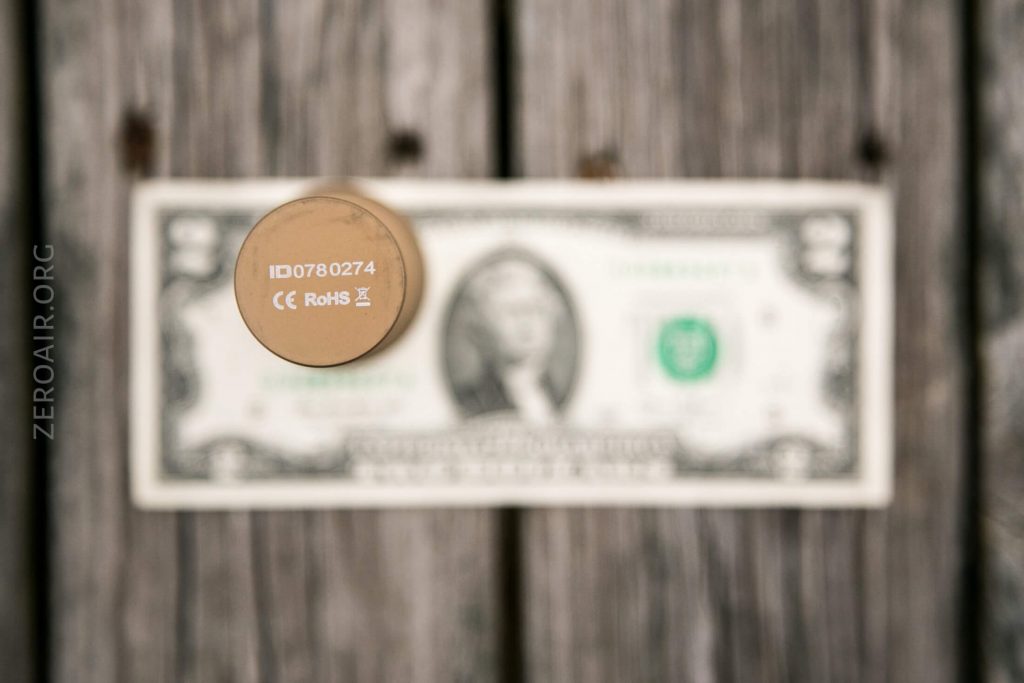 A small round object with markings “ID0780274 CE RoHS” is placed vertically on a blurred U.S. one-dollar bill atop a weathered wooden surface. The website “ZEROAIR.ORG” is visible on the left.