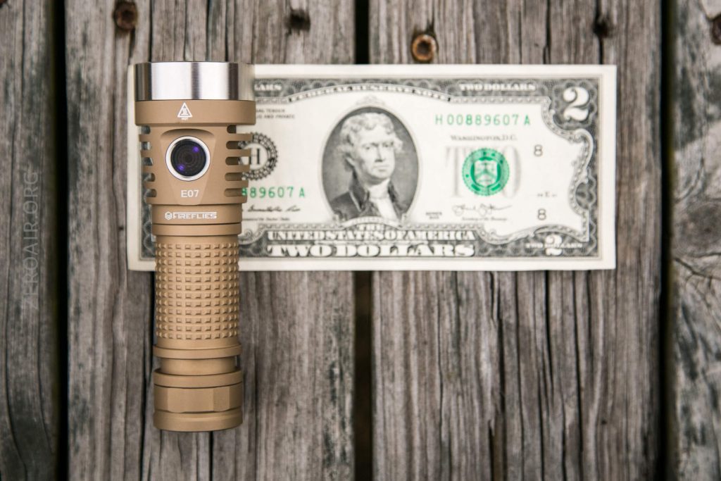 A tan-colored flashlight stands upright in front of a U.S. two-dollar bill, both placed on weathered wooden boards. The flashlight is shorter than the length of the bill.