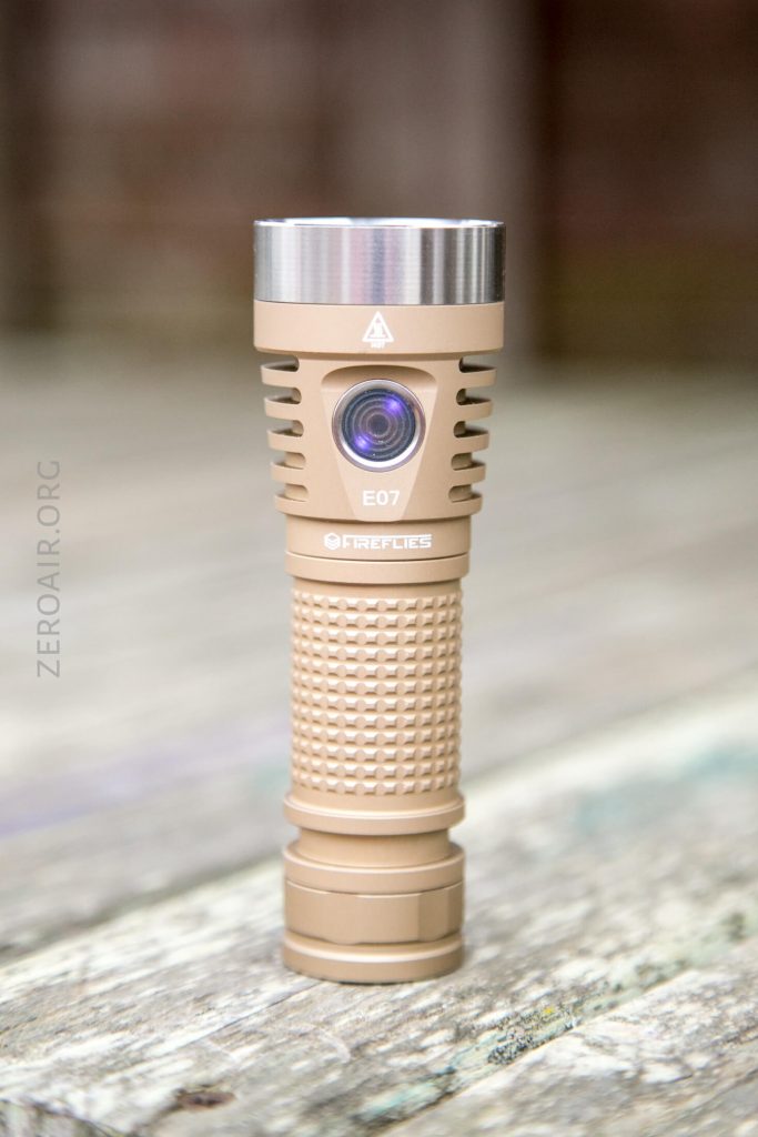 A tan-colored Fireflies E07 flashlight stands upright on a weathered wooden surface, with a metallic silver bezel and textured grip. The lens and product branding are visible.