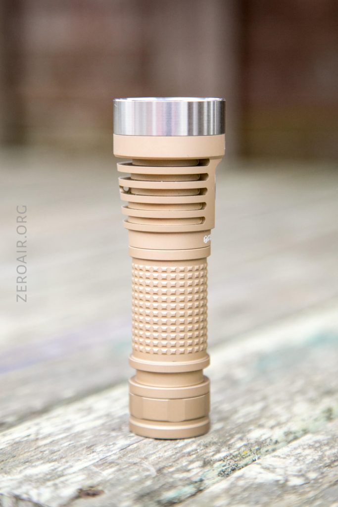 A tan-colored, textured flashlight with a metallic silver top is standing upright on a weathered wooden surface. The background is blurred.