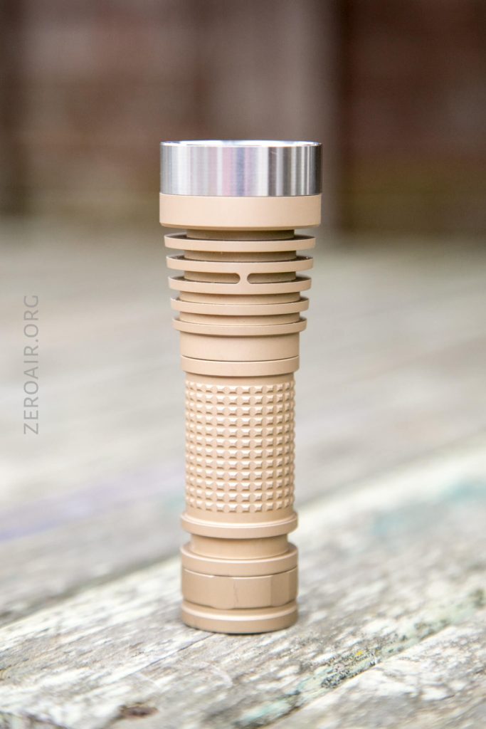 A tan-colored, textured flashlight with a metallic silver head stands upright on a weathered wooden surface. The background is blurred.