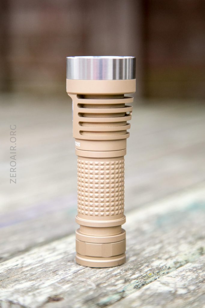 A tan-colored flashlight with a metallic silver head is standing upright on a weathered wooden surface. The flashlight has a textured grip and horizontal grooves near the top. The background is blurred.