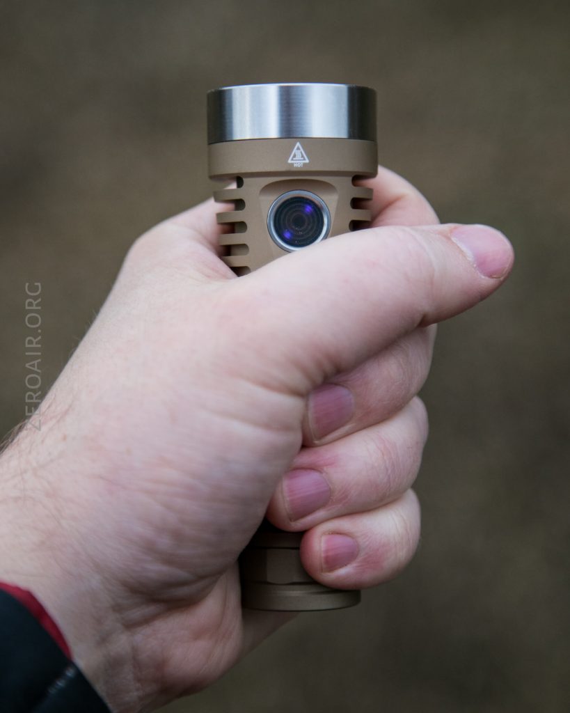 A close-up of a persons hand holding a tan-colored flashlight with a metallic silver top, featuring a visible lens and a warning triangle symbol near the front. The background is blurred.