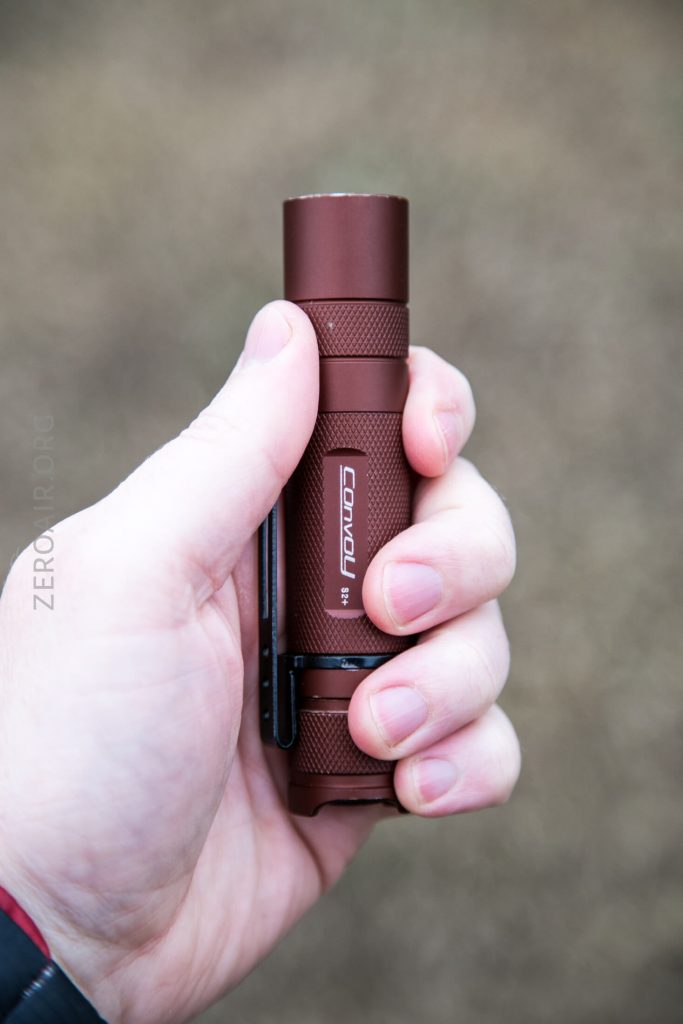 A person’s hand holding a compact, cylindrical, brown Convoy flashlight with a textured grip and pocket clip, against a blurred, neutral outdoor background.