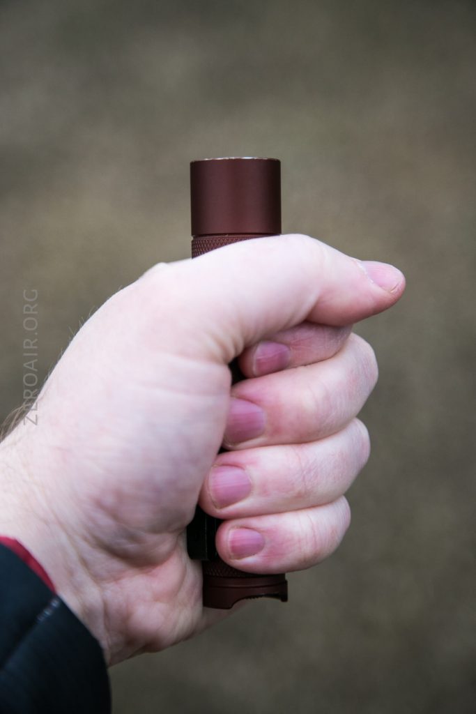 A person holding a small, cylindrical, dark red flashlight in their right hand against a blurred brown background.