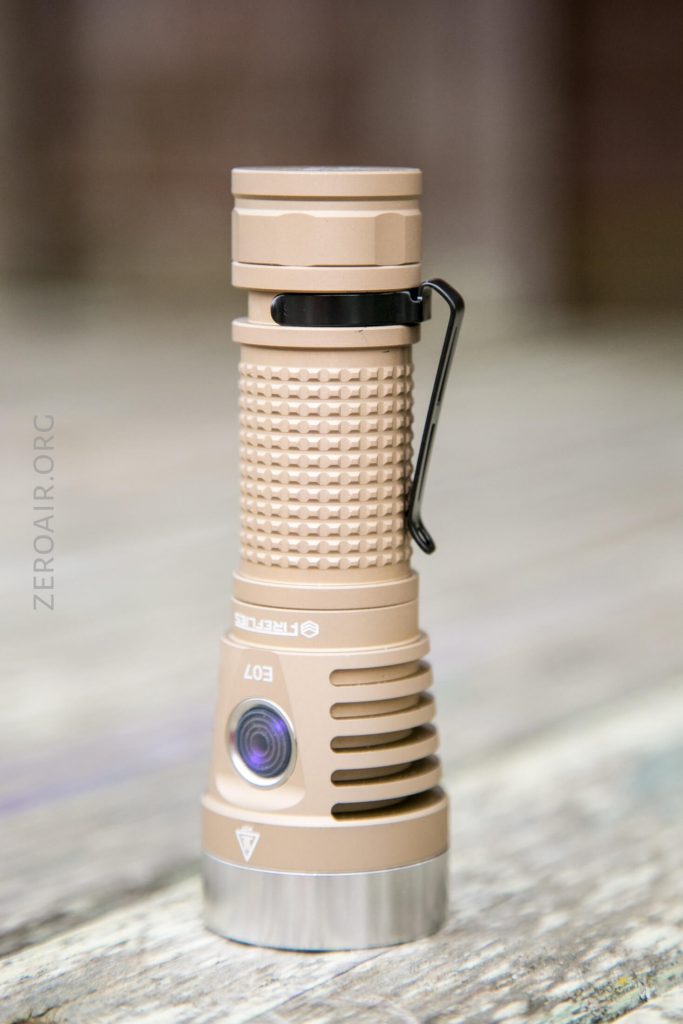 A tan-colored flashlight with a textured grip stands upright on a wooden surface. It has a black pocket clip and a silver button near the head. The background is blurred.
