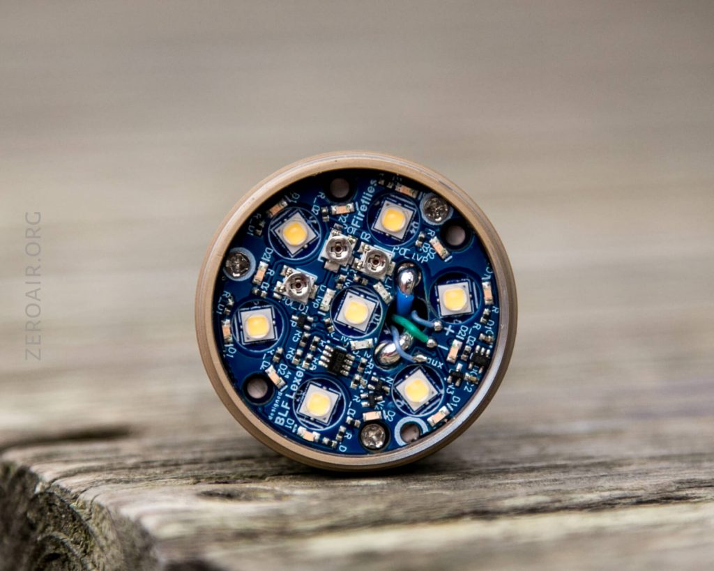 Close-up of a round electronic circuit board featuring multiple LED lights, resistors, and wiring, resting upright on a wooden surface. The text ZEROAIR.ORG is faintly visible along the left edge.