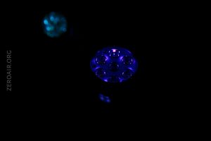 A circular object with multiple small lights emits a blue glow in a dark environment. A second faint blue object appears in the background. The text ZEROAIR.ORG is visible along the left side.