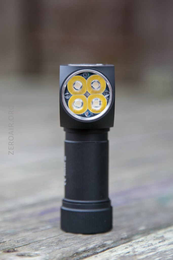 A black handheld flashlight with four yellow LED bulbs arranged in a square pattern stands upright on a wooden surface.