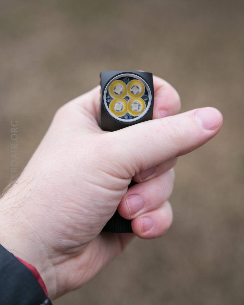 A person holds a black flashlight with four LED bulbs facing the camera. The persons thumb is on top, and the background is blurred.