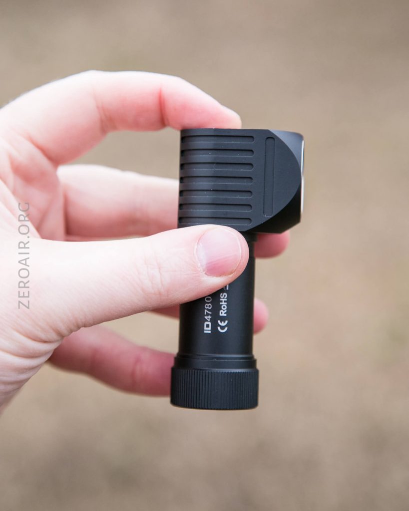A hand holding a small black flashlight with a right-angle head against a blurred brown background. The flashlight has ridged grip details and visible branding and certification marks on the side.