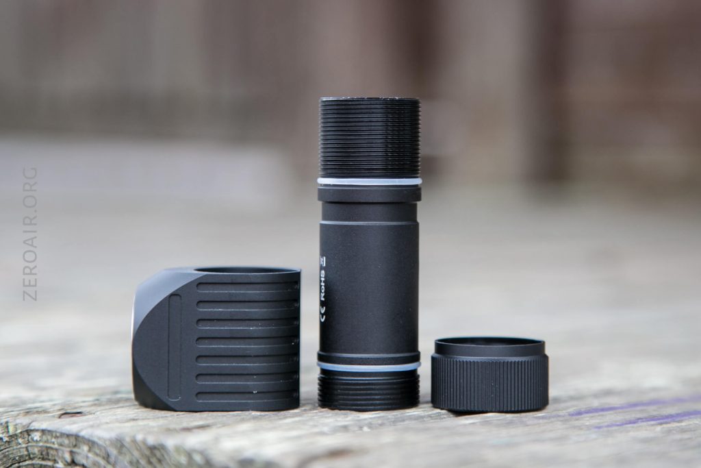 A black cylindrical flashlight is shown disassembled into three parts: the main body, the head, and the tail cap. The components are placed upright on a wooden surface with a blurred background.