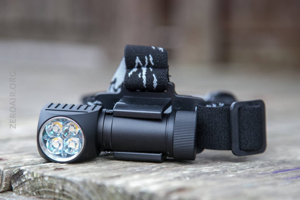 A black headlamp with four LED bulbs and an adjustable head strap rests on a wooden surface. The headlamp has a cylindrical battery compartment and a side switch for operation.