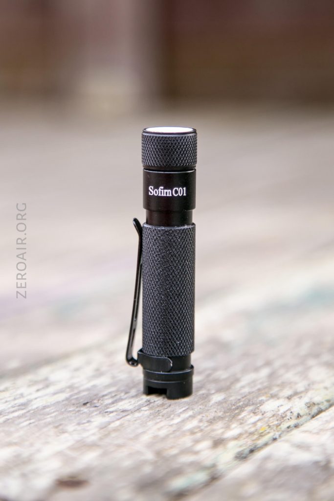 A black Sofirn C01 flashlight stands upright on a wooden surface. The flashlight has a textured grip, a pocket clip, and the product name is printed near the top.