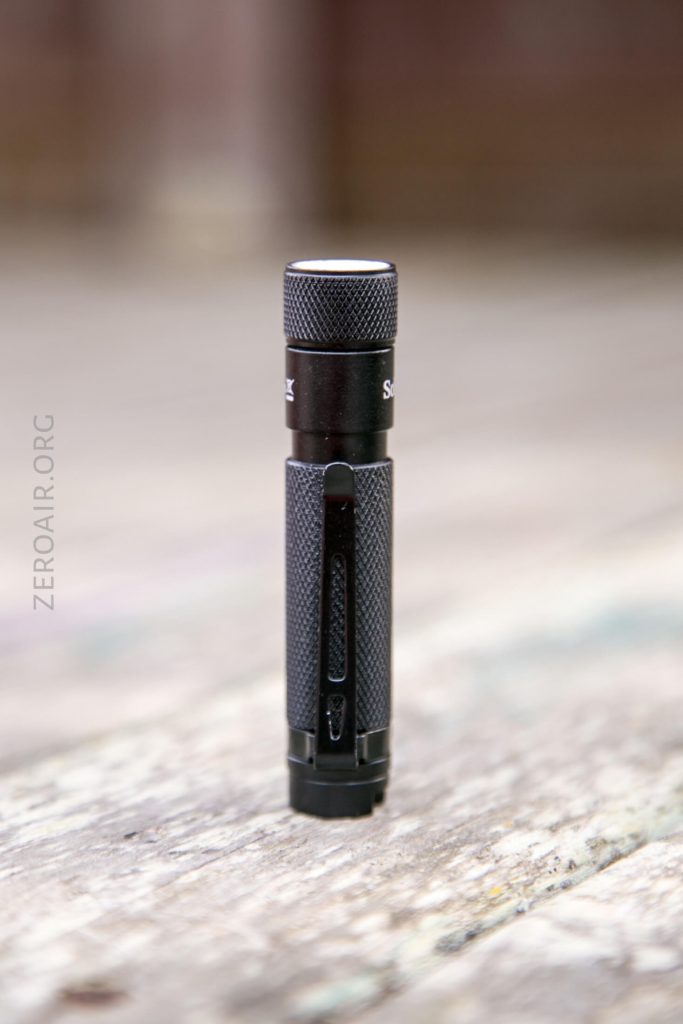 A small, black, textured flashlight standing upright on a weathered wooden surface with a blurred background. The word zeroair.org is faintly visible on the left side of the image.