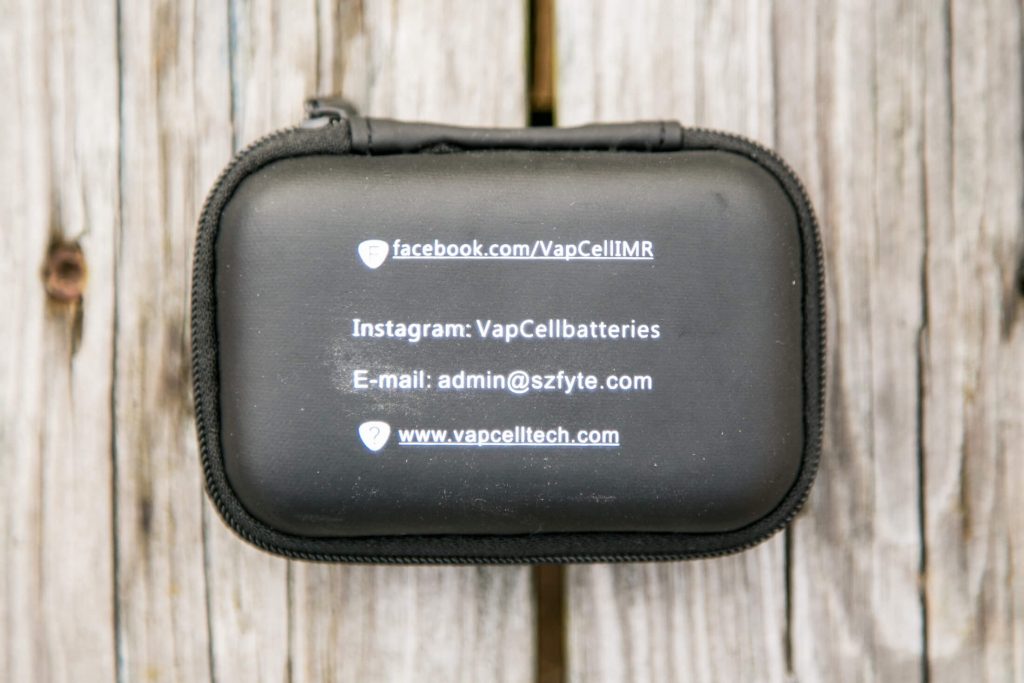 A black zippered case sits on a wooden surface. The case displays social media handles, an email address, and a website for VapCell, a battery company.