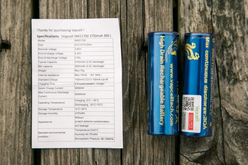 A paper specification sheet and two blue cylindrical Vapcell high drain rechargeable batteries are placed side by side on a wooden surface. The sheet lists technical details for the Vapcell INR21700 battery model.
