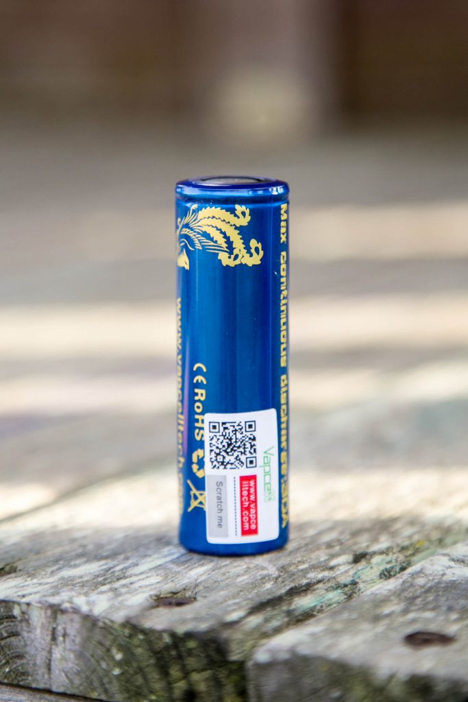 A blue cylindrical battery with gold text and a gold dragon design stands upright on a wooden surface. A QR code and certification labels are visible on the side of the battery. The background is out of focus.