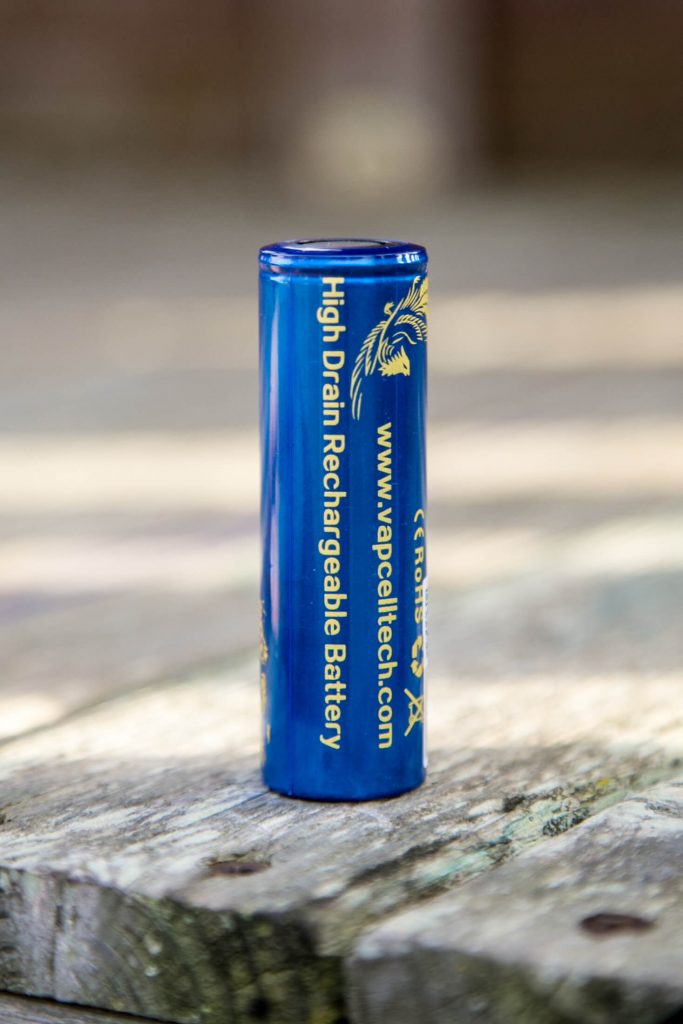 A blue cylindrical battery labeled High Drain Rechargeable Battery stands upright on a weathered wooden surface. The website www.vapcelltech.com is printed vertically on the battery.