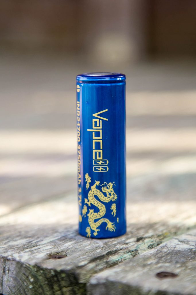 A blue cylindrical battery stands upright on a surface. The battery has gold text that reads Vapcell and features a gold dragon design. The background is blurred.