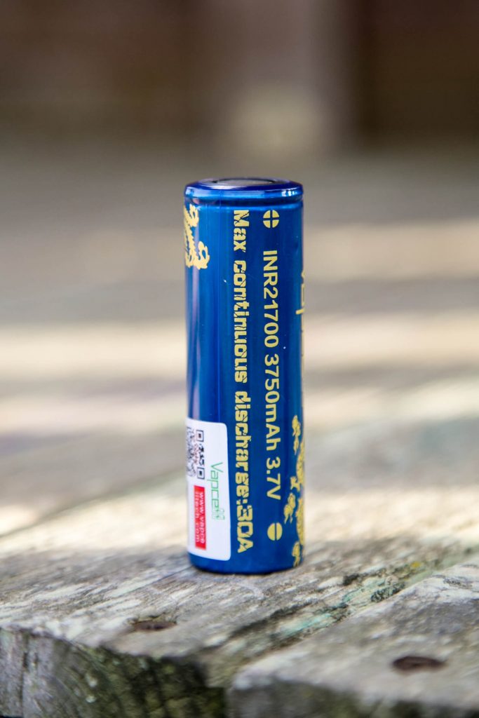 A blue cylindrical battery labeled INR21700, 3750mAh, 3.7V stands upright on a wooden surface. The label also includes Max continuous discharge: 30A and a small QR code.