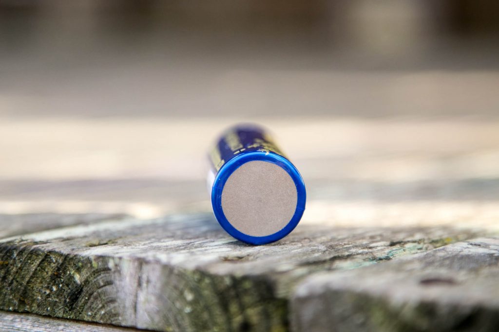 A close-up of a blue cylindrical battery lying on its side on a wooden surface, with the flat, silver-colored terminal facing the camera. The background is out of focus.
