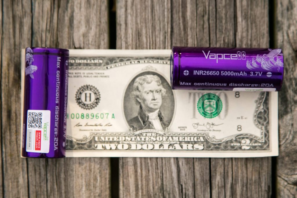 A two-dollar bill lies flat on a wooden surface with two purple Vapcell INR26650 batteries, one on top and one to the right of the bill. The batteries are labeled 5000mAh, 3.7V, and 20A max discharge.