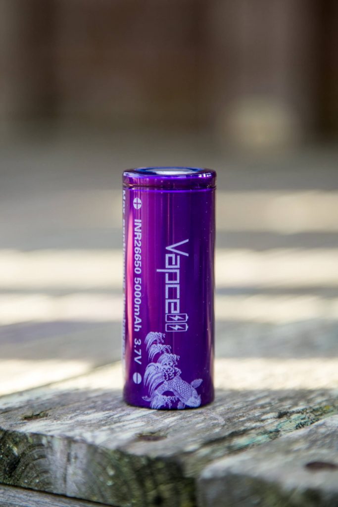 A purple cylindrical battery labeled VAPCELL INR26650 5000mAh 3.7V stands upright on a wooden surface, with a blurred background.