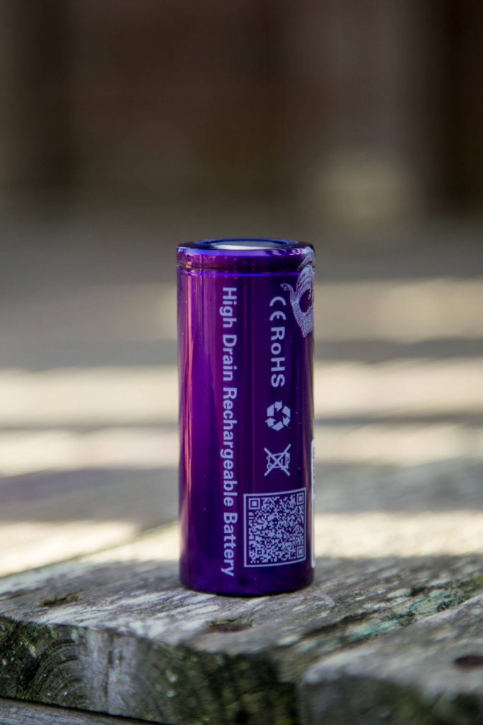A purple cylindrical high drain rechargeable battery stands upright on a weathered wooden surface. The battery has visible safety symbols, a QR code, and text including “High Drain Rechargeable Battery.”