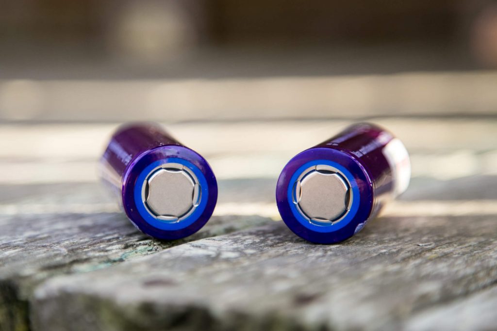 Two cylindrical batteries with purple casings and blue rims are lying side by side on a wooden surface, viewed from the terminal end.