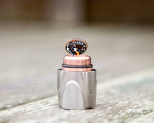 A close-up of a cylindrical metal object with a copper component and visible electronic circuitry on top, standing upright on a wooden surface.