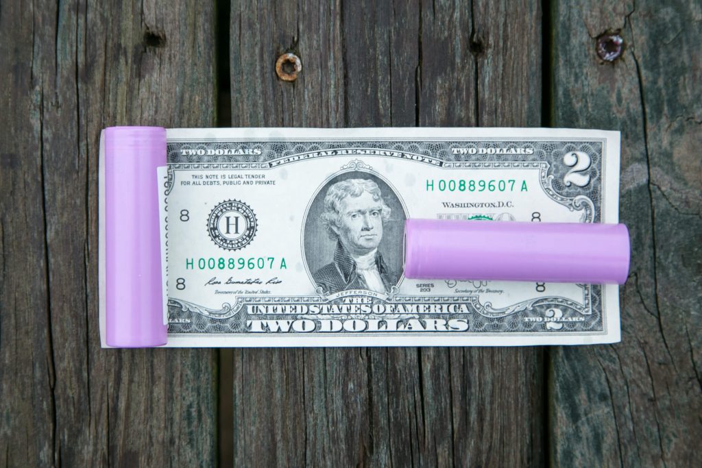 A two-dollar bill lies on a wooden surface with two purple cylindrical objects, one on each side of the bill, partially covering its edges.