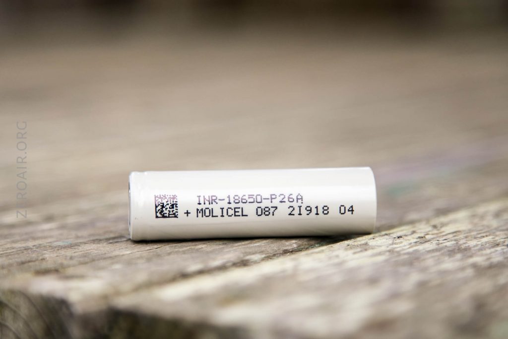 A white cylindrical battery labeled INR-18650-P26A + MOLICEL 087 21910 04 rests on a wooden surface. The battery has printed text and a small QR code.