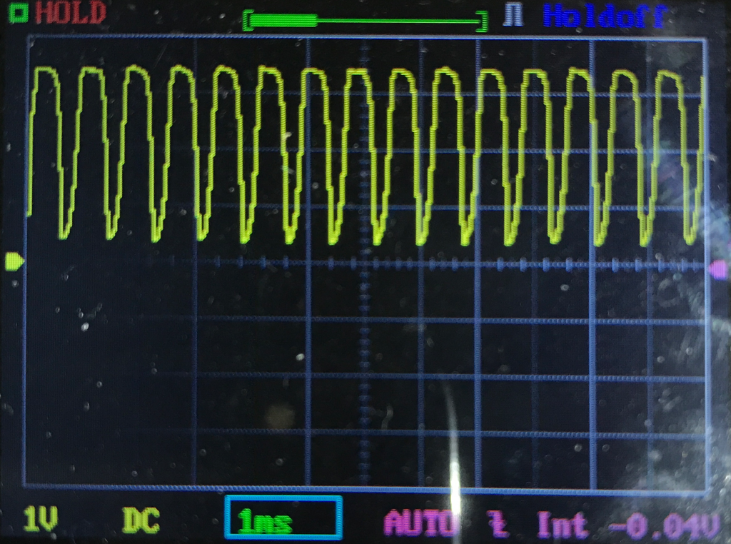 Oscilloscope display showing a yellow square waveform signal with a periodic pattern, grid lines in the background, and measurement settings along the bottom edge of the screen.