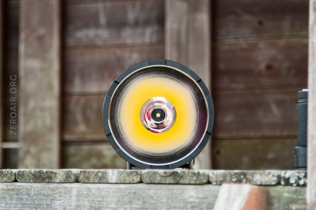 A close-up view of a modern round LED light or spotlight with a reflective surface, a central black circle, and a yellow ring, positioned against a blurred wooden background.