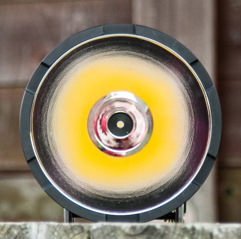 A close-up view of a modern round LED light or spotlight with a reflective surface, a central black circle, and a yellow ring, positioned against a blurred wooden background.