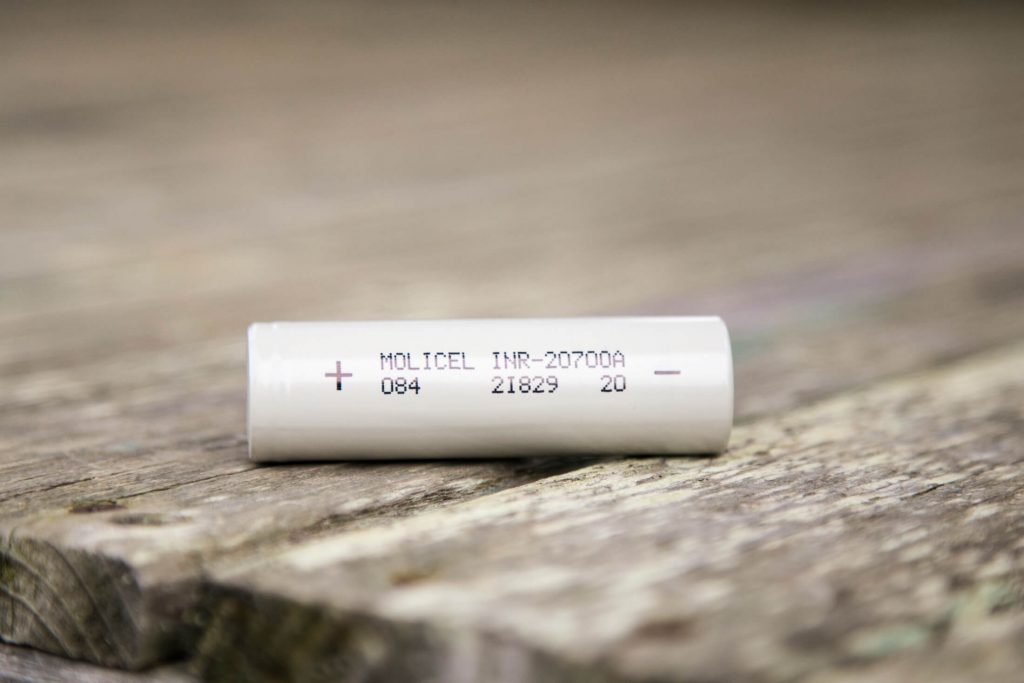 A white cylindrical battery labeled “Molicel INR-20700A” rests on a weathered wooden surface. The battery shows a plus and minus sign and additional numbers printed on its side.