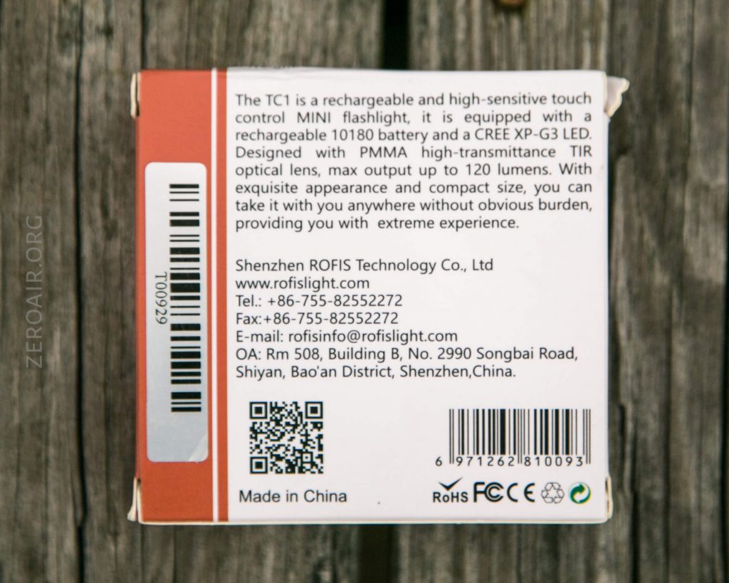 A product box displays specifications for the TC1 MINI flashlight, including details about battery, LED, output, company contact information, barcodes, and certification symbols, placed on a wooden surface.