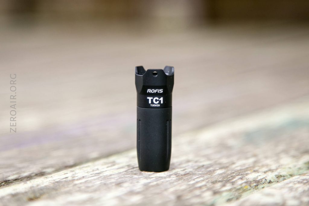 A small black flashlight labeled ROFIS TC1 stands upright on a wooden surface, with the background softly blurred.