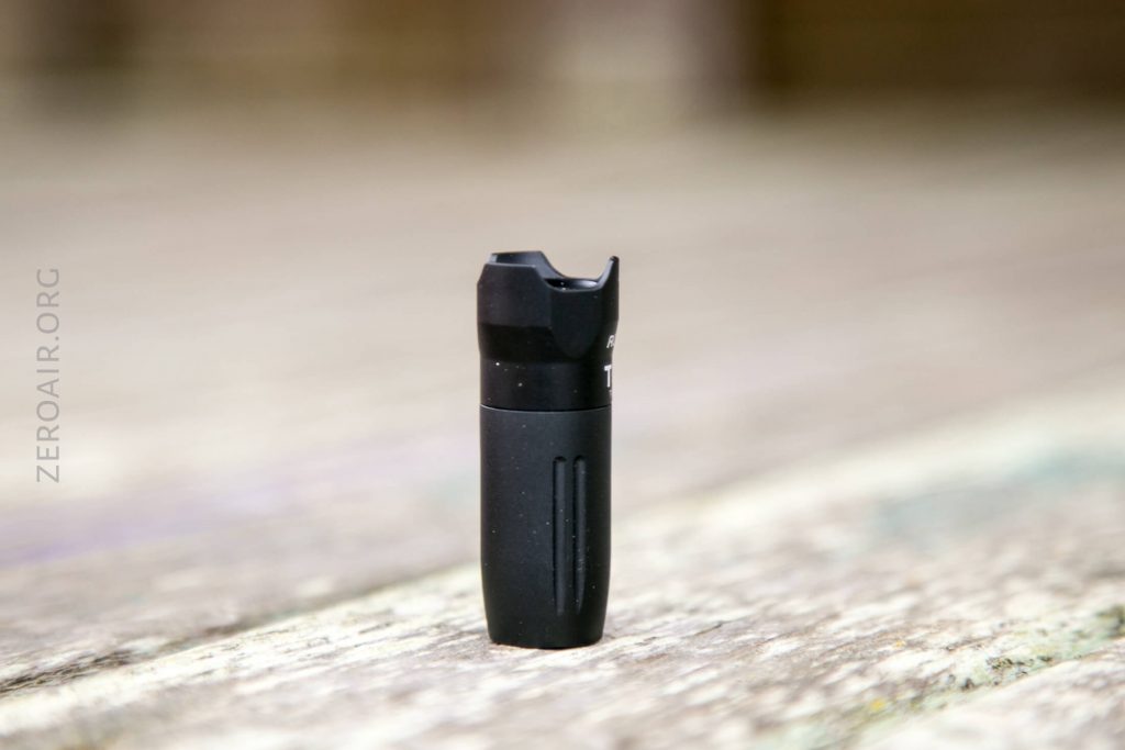 A small, black cylindrical object with a notched top stands upright on a wooden surface. The background is out of focus and the branding “ZEROAIR.ORG” is faintly visible along the left side.