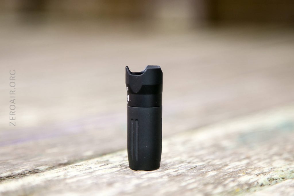 A small, black, cylindrical object with a notched top stands upright on a wooden surface. The background is blurred, and the object has vertical grooves near the bottom. The text ZEROAIR.ORG is visible on the left side.