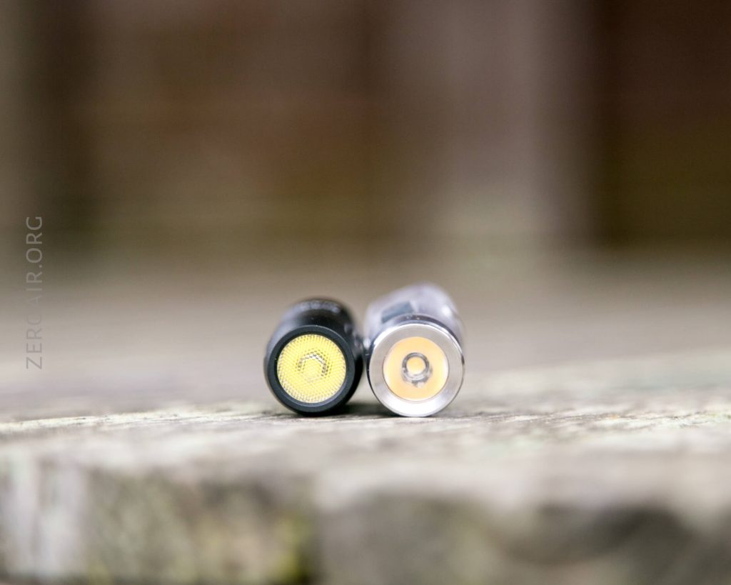 Two vape cartridges are lying horizontally on a textured surface, shown from the end with the mouthpiece on the left and the metal contact side on the right. The background is blurred.