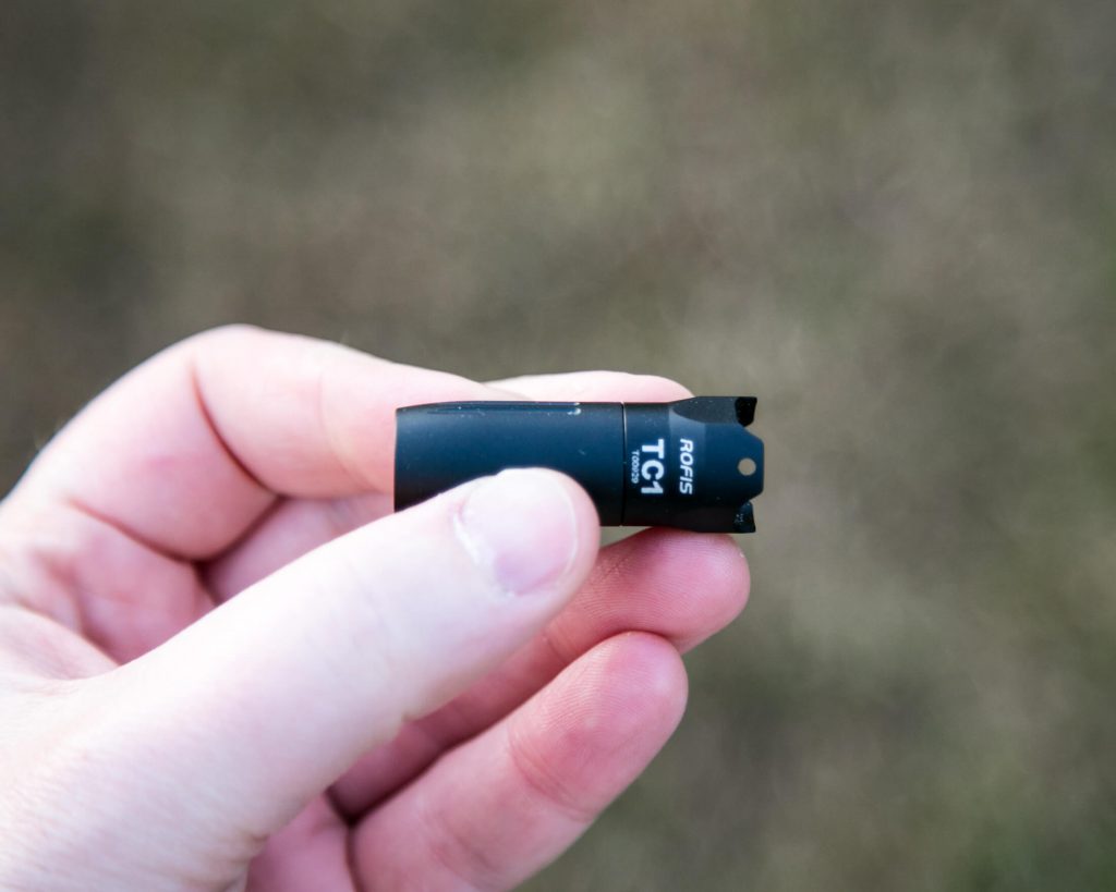 A person holds a small, black, cylindrical object with white text reading “TC5” and “noris” against a blurred outdoor background.
