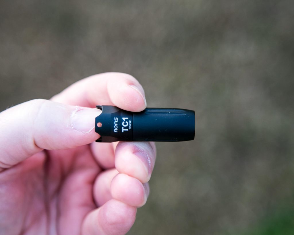 A left hand holds a small black ROFIS TC1 flashlight outdoors. The flashlight is cylindrical and compact, with product marking visible. The background is blurred grass or foliage.