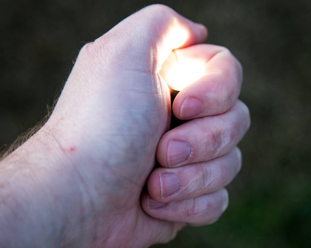 A close-up of a persons hand holding and activating a lighter, producing a small flame. The background is blurred and out of focus.