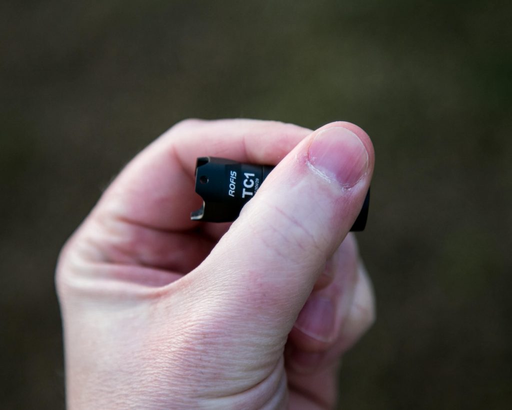A close-up of a persons hand holding a small black electronic component labeled TC1 RG45 between their thumb and index finger, with a blurred background.